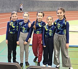 Fünf junge Turnerinnen der SG Bruchköbel in blau-gelben Turnanzügen lächeln gemeinsam in einer Leichtathletikhalle.“