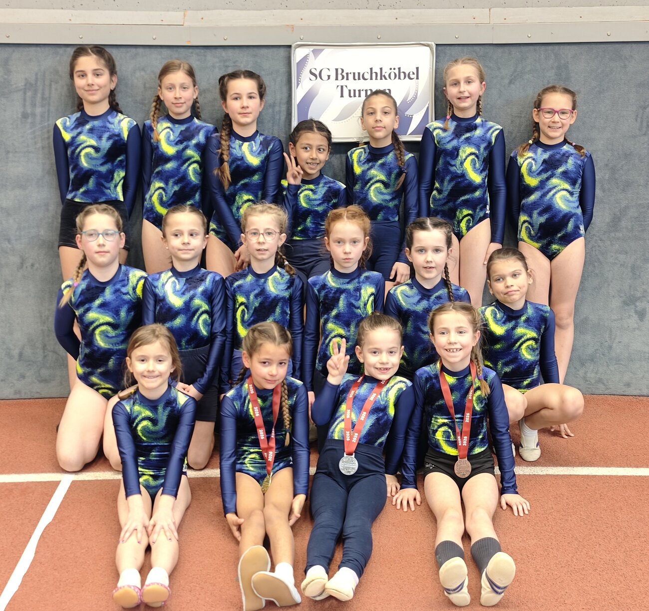 Gruppenfoto von 16 jungen Turnerinnen der SG Bruchköbel in blau-gelben Turnanzügen, einige tragen Medaillen, vor einem Vereinsschild.“
