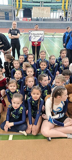 Fünf junge Turnerinnen der SG Bruchköbel in blau-gelben Turnanzügen lächeln gemeinsam in einer Leichtathletikhalle.